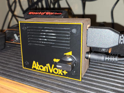 A small box with a black plastic front and wood sides. The front is trimmed in orange and has a speaker grill at the top. A knob is currently turned to 'off'. The word 'AtariVox+' is emblazoned on it in orange. Two cables come out of the right side.