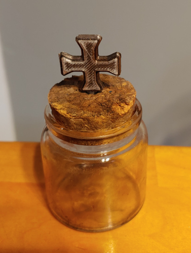 A small, weathered, glass, bottle with a cork. A silver cross sticks out of the top of the cork.