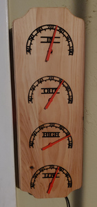 A wooden board hanging upright on a wall. Four semi-circle indicator dials are spread across its length, with red needles pointing to numbers.
