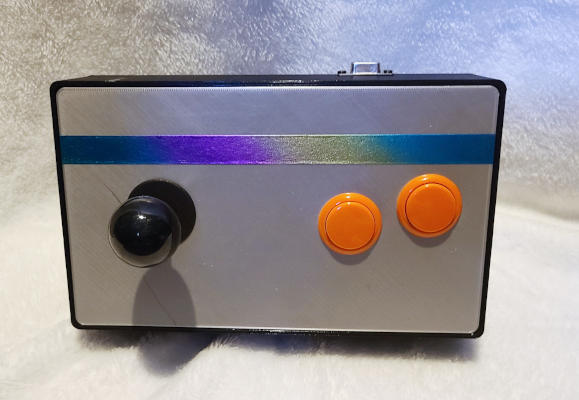 A black box with rounded corners and a silver face. A black ball-top joystick and two orange buttons are installed in the face. There is a rainbow stripe across the top of the face. A connector projects slightly from the back of the box.