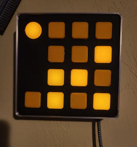 A square object hanging on a wall. It has thin silver sides and a black face. The face is covered in a grid of orange squares, and one white circle. Some of the orange squares are lit up.