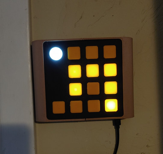 A square object hanging on a wall. It has curved beige sides and a black face. The face is covered in a grid of orange squares, and one white circle. Some of the orange squares are lit up.