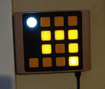 A square object hanging on a wall. It has curved beige sides and a black face. The face is covered in a grid of orange squares, and one white circle. Some of the orange squares are lit up.