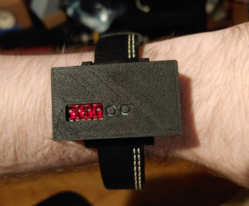 A black plastic box with a leather strap, being worn on a wrist. There are a small red indicator and two buttons on the face of the box.