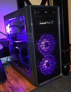 Shiny gray tower PC with a black mesh front. The side is clear showing a dimly lit interior. Fans and a Gigabyte logo glow purple.