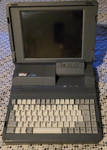 Chunky, dark grey laptop. The screen hinge is offset to the right, the keyboard is full-height with beige and gray keys.