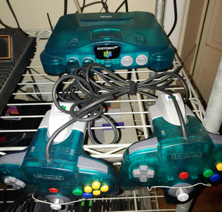 A small, clear, blue-green device with excessive curves, and two matching controllers with multicolored buttons.