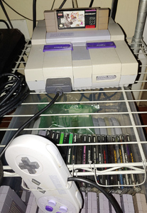 A light gray box, with other box shapes protruding from it and two rectangluar purple switches.
          There is a rounded slab of a controller in front of it with matching purple buttons.
          A darker gray cartridge is sticking out of the top of the console, labeled Chrono Trigger.
          The plastic of the  console and cartridge are somewhat yellowed.