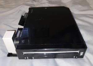 A shiny black slab with a slot for a CD. Flaps along one side are open. A light gray box and a smaller black one stick out of that side.