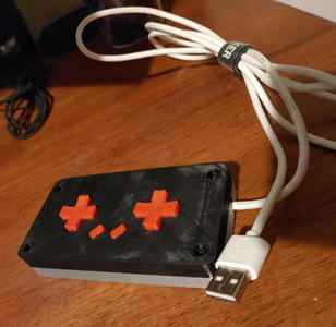 A rectangular gamepad with two cross-shaped directional pads, and two slanted rectangular buttons in the middle. A USB cable comes out of the right side.