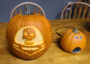 A large pumpkin with an image of a skull in a space helmet carved in relief. A smaller pumpkin next to it has two metal eyes and round speaker in the place of a mouth. A black cable connects the two.