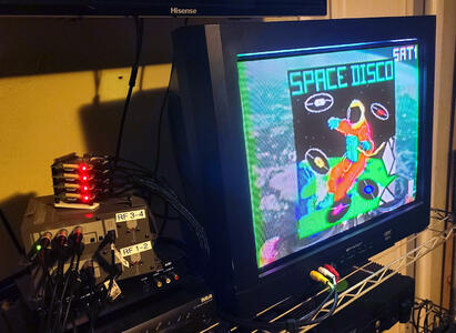 A CRT Television showing a 'Space Disco' program. Next to it is a stack of tiny circuit boards, each with a glowing LED. Underneath the circuit boards are two metal boxes with cables coming out of them, labled 'RF 3-4' and 'RF 1-2'.