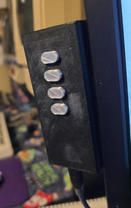 A tall box, about 90mm high, with four capsule-shaped buttons on the front. It is suspended in air by the right side which is attached to the side of a monitor. A USB cable is plugged into the bottom.