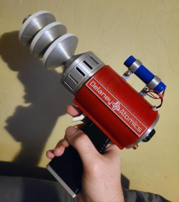 A gun-shaped object with three conical discs on the silver barrel, a red body, a black grip, a cylindrical battery on top, and a dial and button on the back. There is text on the side reading 'Delaney Atomics'.