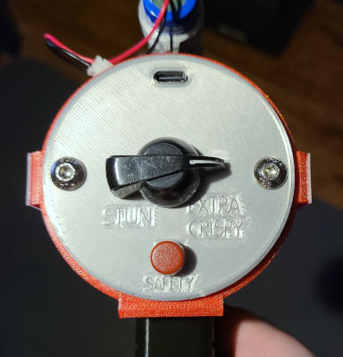A close up of the circular, silver, back panel of the raygun. There is a dial with the settings 'Stun' and 'Extra Crispy'. A red button labeled 'Safety' is below the dial.