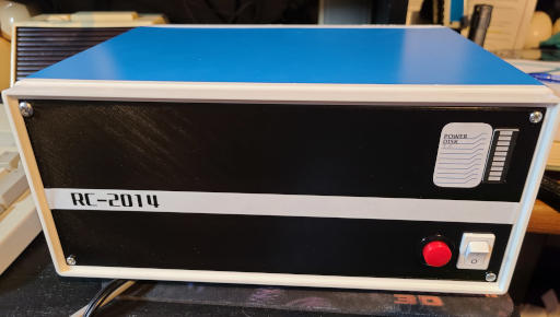 An oblong box with a blue top and a black front, with a white border. A label across the front says 'RC2014'. There is a labeled column of LEDs on the top right. There is a red button and a white switch on the bottom right.