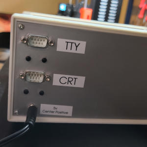 The silver back side of the box, with two d-shaped connectors labeled 'TTY' and 'CRT', and a round connector with a plug in it labeled '5v Center Positive'