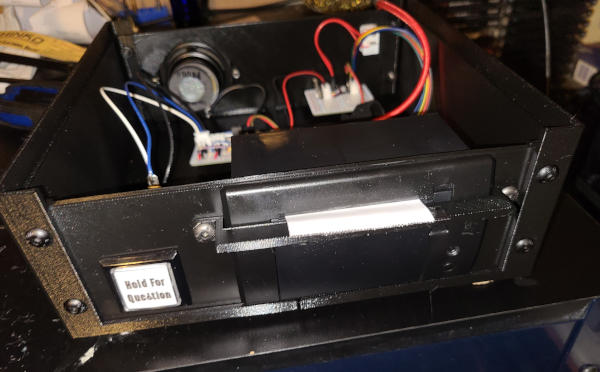 A wide, shallow, black platic box with an open top. A button labeled 'Hold for Question' is on the front, next to a receipt printer. A speaker and switch are attached to the back. Circuit boards and wires are visible inside.
