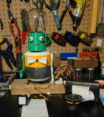 A robot made mainly of mismatched green, yellow, and white plastic. The lower torso has a tarnsihsed silver finish. The robot sits on a piece of wood. 