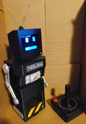 A black toy robot with a skirt-like lower section, thin silver waist, a boxy torso, thin silver arms, silver tubular neck, and an old-fashioned TV with rabbit-ear antenna as a head. An Atari joystick sits next to it.