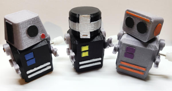 Three small robots, each with head, boxy torso, arms, winding cranks, and wheels. They are mostly black and silver, with painted details in other colors. Each head is a different shape.