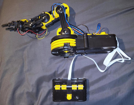 An industrial-style robot arm built of black and yellow plastic. A flat, gray cable runs to a remote control.