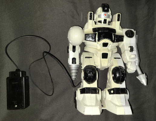A white plastic robot with black and silver accents. It has legs with large feet, and one arm ends in a conical drill. The head is reminiscent of jet pilot helmet, but with a red LED in the middle of the visor. A wire leads to a separate black box.
