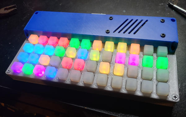 A rectangular plastic slab. There is a raised blue section along the back with a rocker switch and a speaker grill. The lower section is silver with a 12x4 grid of light-up square buttons.