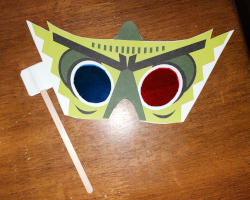 A paper mask that covers the area around the eyes. It is printed with  a green monster design. The left eye has a red lens and the right eye has a blue lens. There is a thin wooden handle on one side.