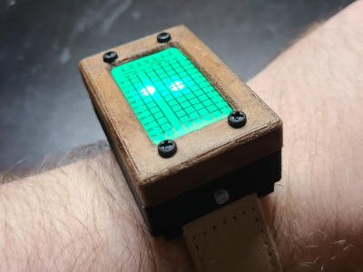 A rectangular watch with a wood bezel. The face is green with a black grid. Two points on the grid are lit up.