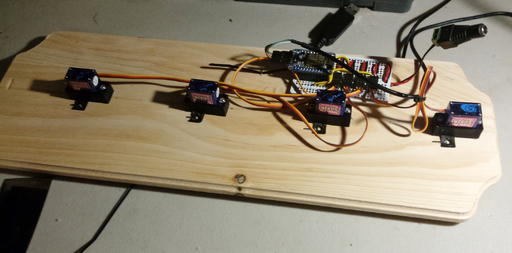 The same wooden board, with small blue servo motor attached at intevals. A circuit board is attached next to one of them.