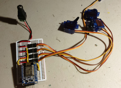 A small blue circuit board attached to a large white prototyping board. Pieces of wire connect different points on the prototyping board, and four cables snake off the board to small blue servo motors.