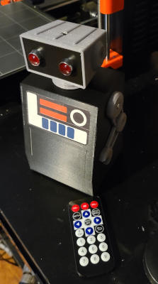 A toy robot with a black trapezoidal body, small silver arms, a silver trapezoidal head, and a black face with two large red LED eyes.