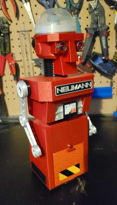 A red toy robot with a skirt-like lower section, thin black waist, a boxy torso, thin silver arms, black tubular neck, and an angular sculpted head. The head has two large red LED eyes and a clear dome cranium.